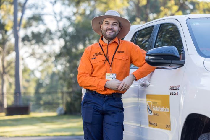 A male staff member wearing a bright orange long sleeve shirt and government ID card. He is standing in front of a fire ant program vehicle. The program's logo is on the door of the vehicle.